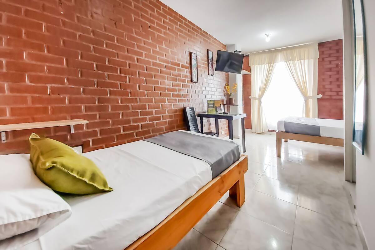 A room with a brick wall, a bed with a pillow, a small table with a TV, and a bench in the background.