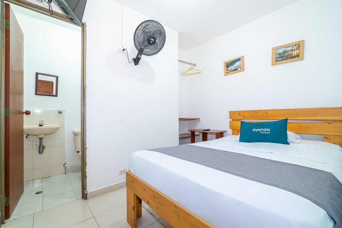 A bedroom with a bed, a bedside table, a wall-mounted fan, a hanging light, and a small wooden table.