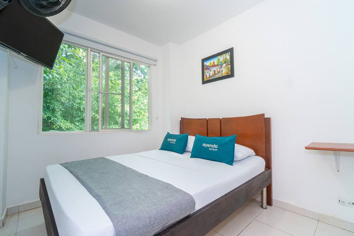 A neatly made bed with a grey blanket and two teal pillows, a wooden headboard, a window with a view of greenery, and a small wooden shelf.