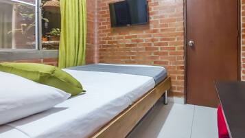 A room with a bed, a TV mounted on the brick wall, a wooden desk, and a window with curtains.