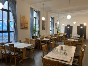 A restaurant with multiple tables and chairs, large windows with curtains, and hanging pendant lights.