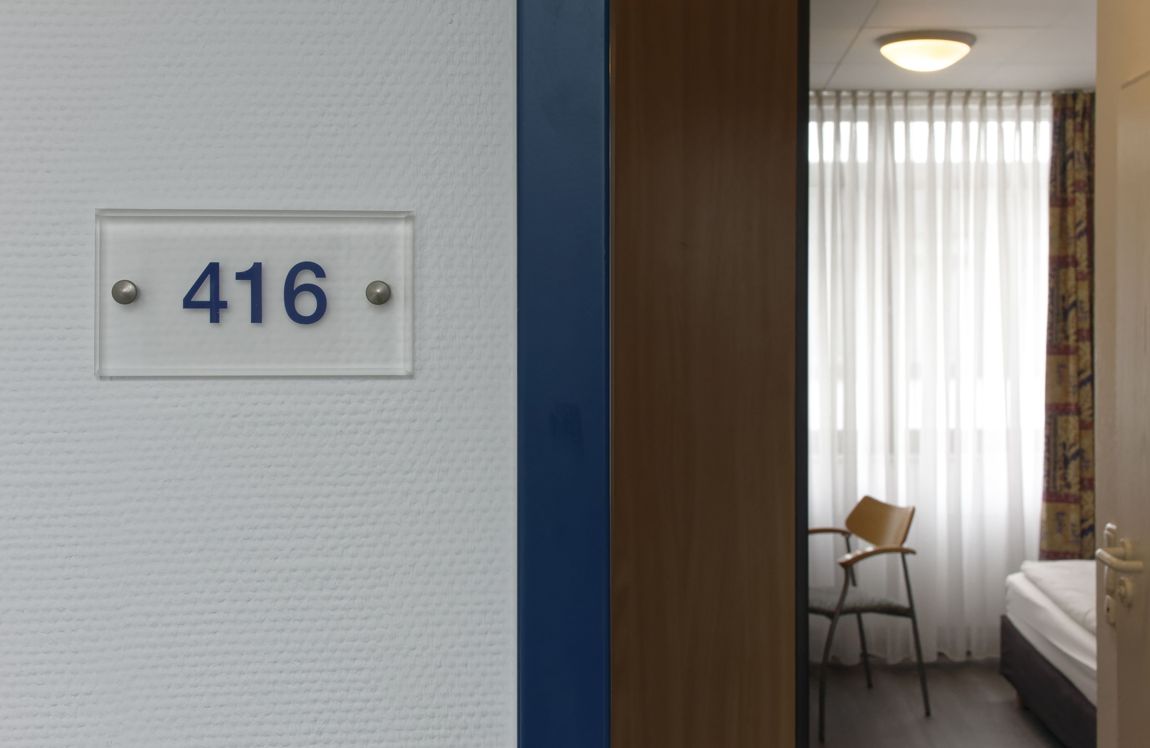 A room with a number sign '416' on the wall, a chair, a bed, and a window with curtains.