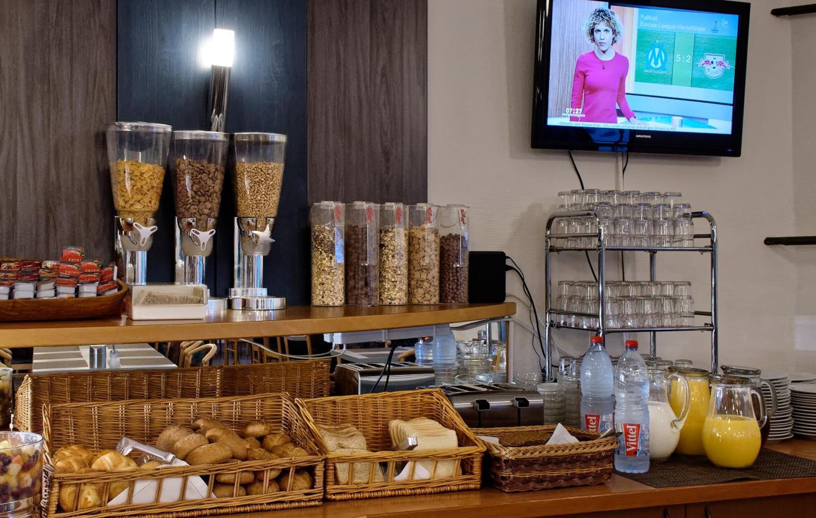 En morgenmadsbuffet med forskellige retter og drikkevarer, et fjernsyn der viser nyhedsudsendelse og et glasdisplay.