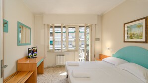 A bedroom with a bed, a wooden nightstand, a desk, a mirror, and a window with a view of buildings. - Hotel Stella d'Italia (Viareggio)
