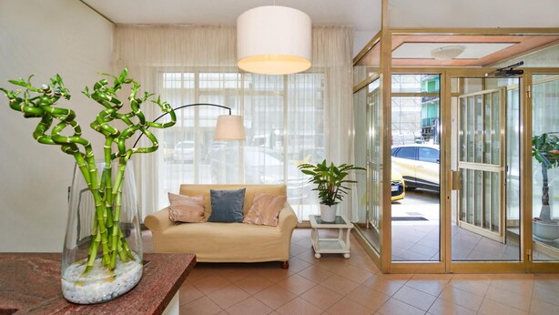 A modern living room with a sofa, a glass vase with green plants, a hanging light fixture, and a view through a glass door to the outside. - Hotel Stella d'Italia (Viareggio)