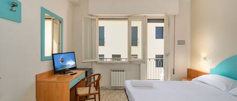 Habitación de hotel con cama, escritorio con computadora, silla, ventana con vista a edificios y radiador.