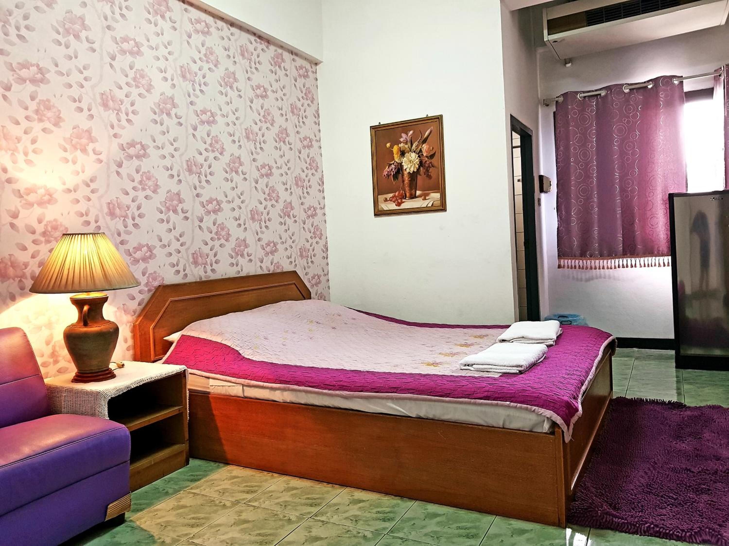Une chambre à coucher avec un lit en bois, un fauteuil violet, une table de chevet avec une lampe, un papier peint à motifs floraux et un tableau accroché au mur.