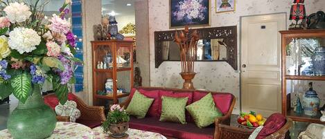 Una sala de estar acogedora con un sofá, sillas de mimbre, una mesa con mantel floreado y un jarrón grande de flores.