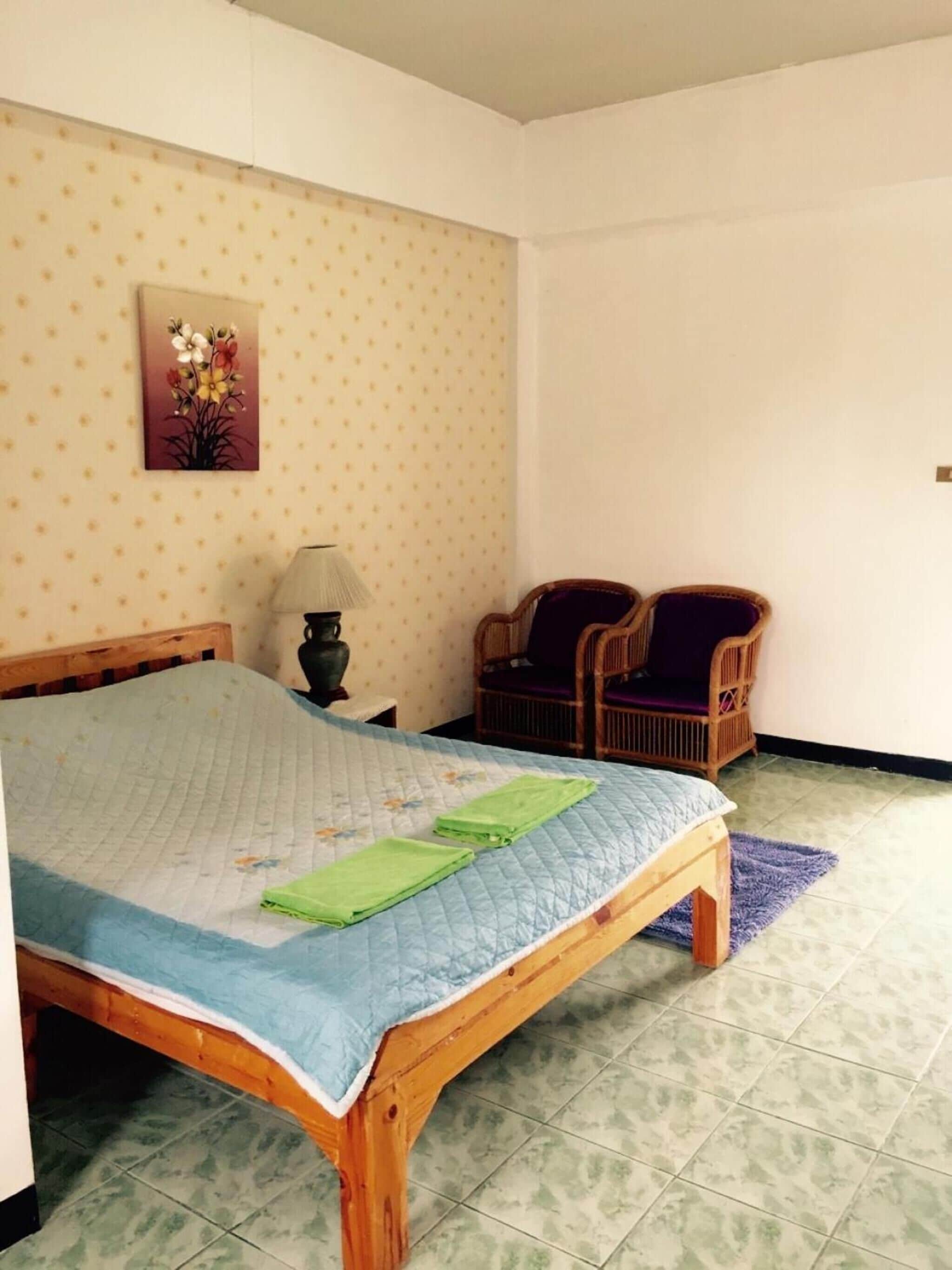 A bedroom with a wooden bed, a nightstand, two wicker chairs, a wall-mounted lamp, and a floral painting.