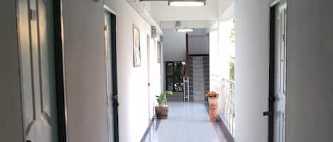 A long, narrow hallway with tiled floors, doors on both sides, and potted plants along the walls.