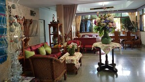 A spacious living room with a red sofa, wicker chairs, a dining table with floral tablecloths, and a large vase of flowers. - Wiang Inn Mansion (Chiang Mai)