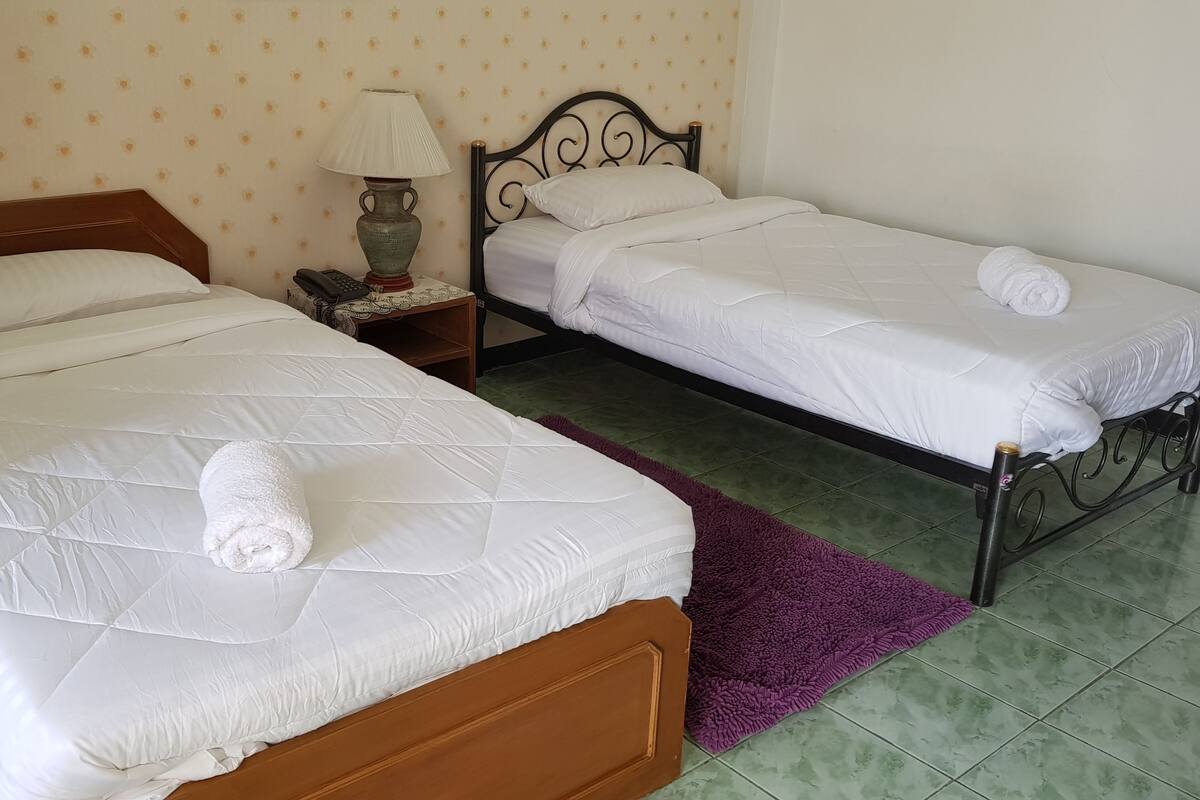 Two single beds with white linens and rolled towels, a nightstand with a lamp, and a purple rug on a tiled floor.