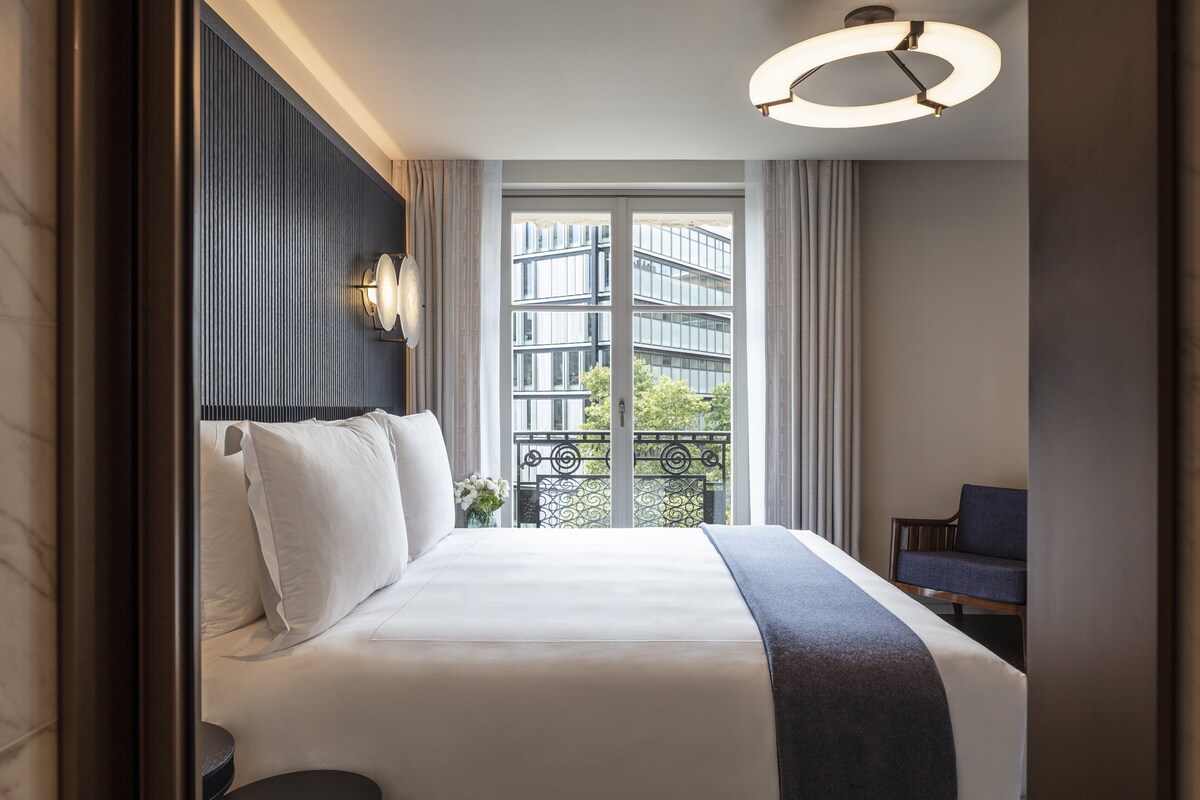 Ett hotellrum med en stor säng, utsikt över en byggnad genom ett fönster, en stol och en modern lampa.