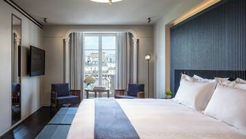 Une chambre d’hôtel avec un grand lit, deux fauteuils, une petite table sur laquelle est posé un vase de fleurs, un téléviseur à écran plat et une vue sur les bâtiments par une grande fenêtre.