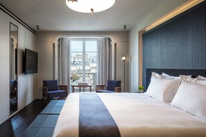 A hotel room with a large bed, two armchairs, a small table with a vase of flowers, a flat-screen TV, and a view of buildings through a large window.