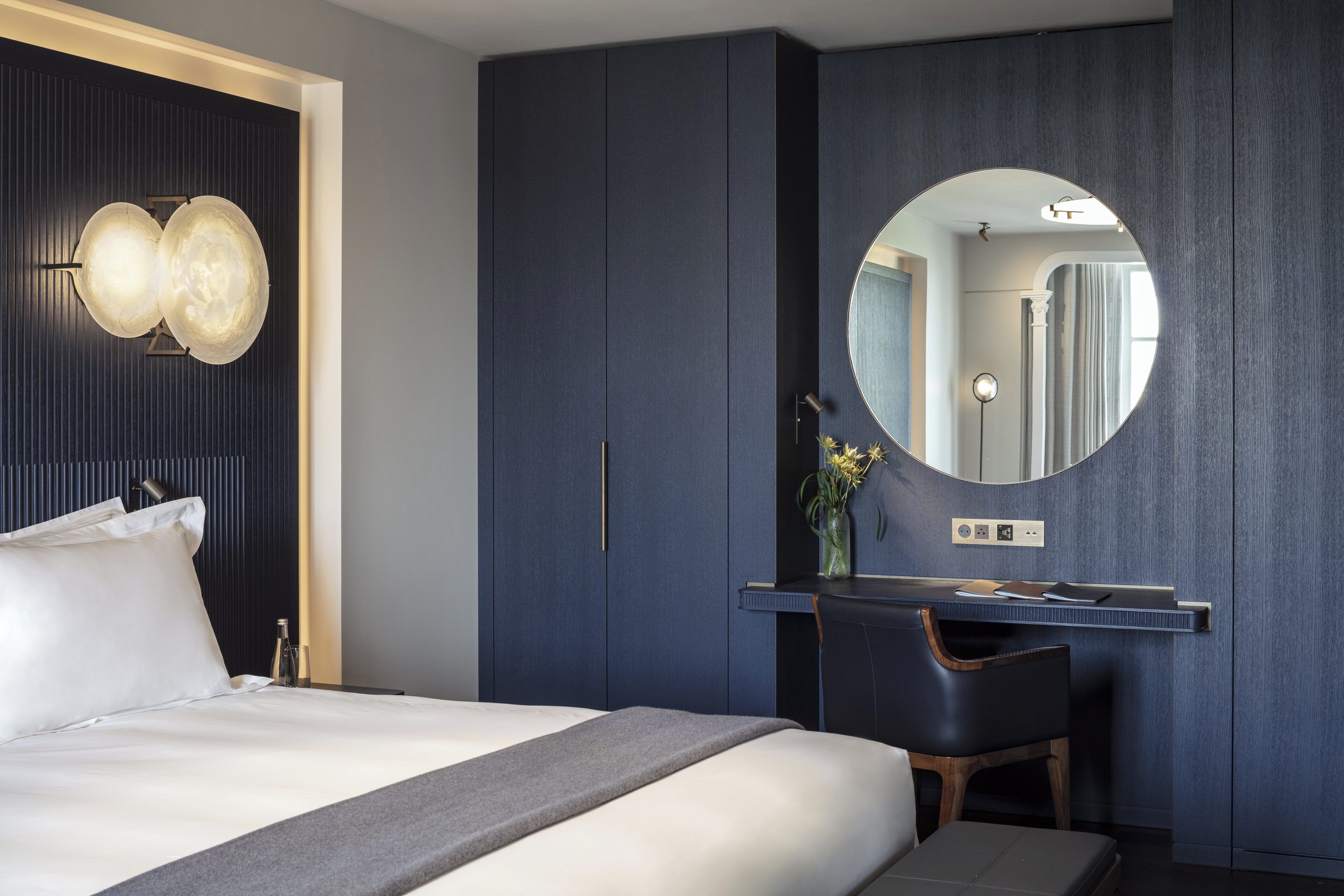 A modern bedroom with a bed, a desk, a chair, a mirror, and a wall-mounted light fixture.