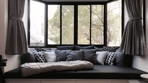 A cozy seating area with a built-in bench, grey cushions, and a view of trees through a large window with black frames. - Stay in Style The Heart of Nimman R208 (Chiang Mai)