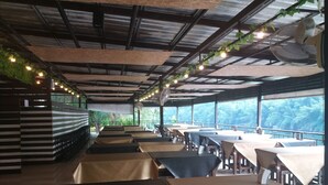 A restaurant with multiple tables and chairs, a high ceiling with exposed beams, and large windows offering a view of the outdoors. - Ingphupha Resort and Restaurant (Si Sawat)