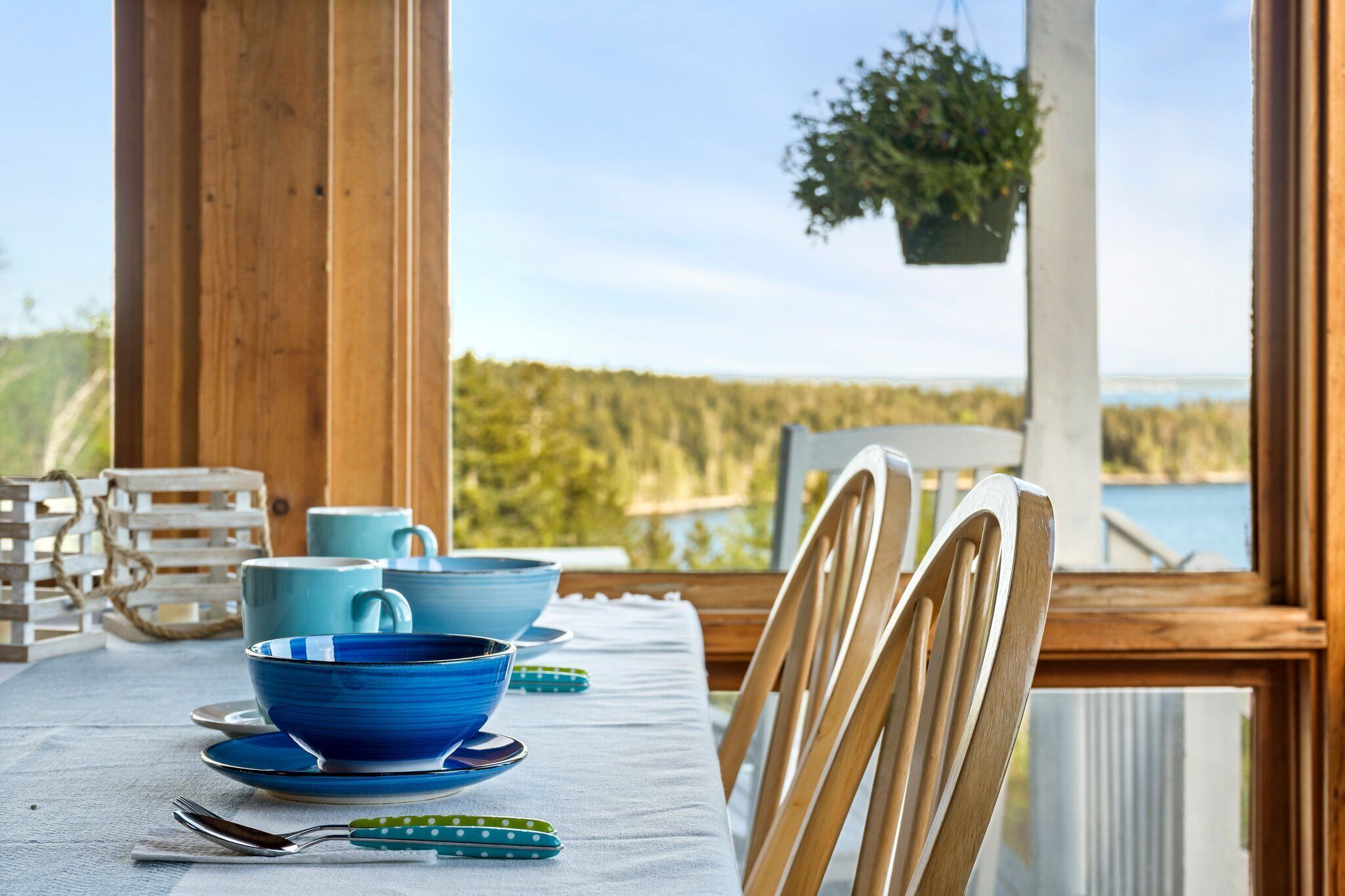 A dining table set with blue bowls and plates, wooden chairs, and a view of a lake through large windows.