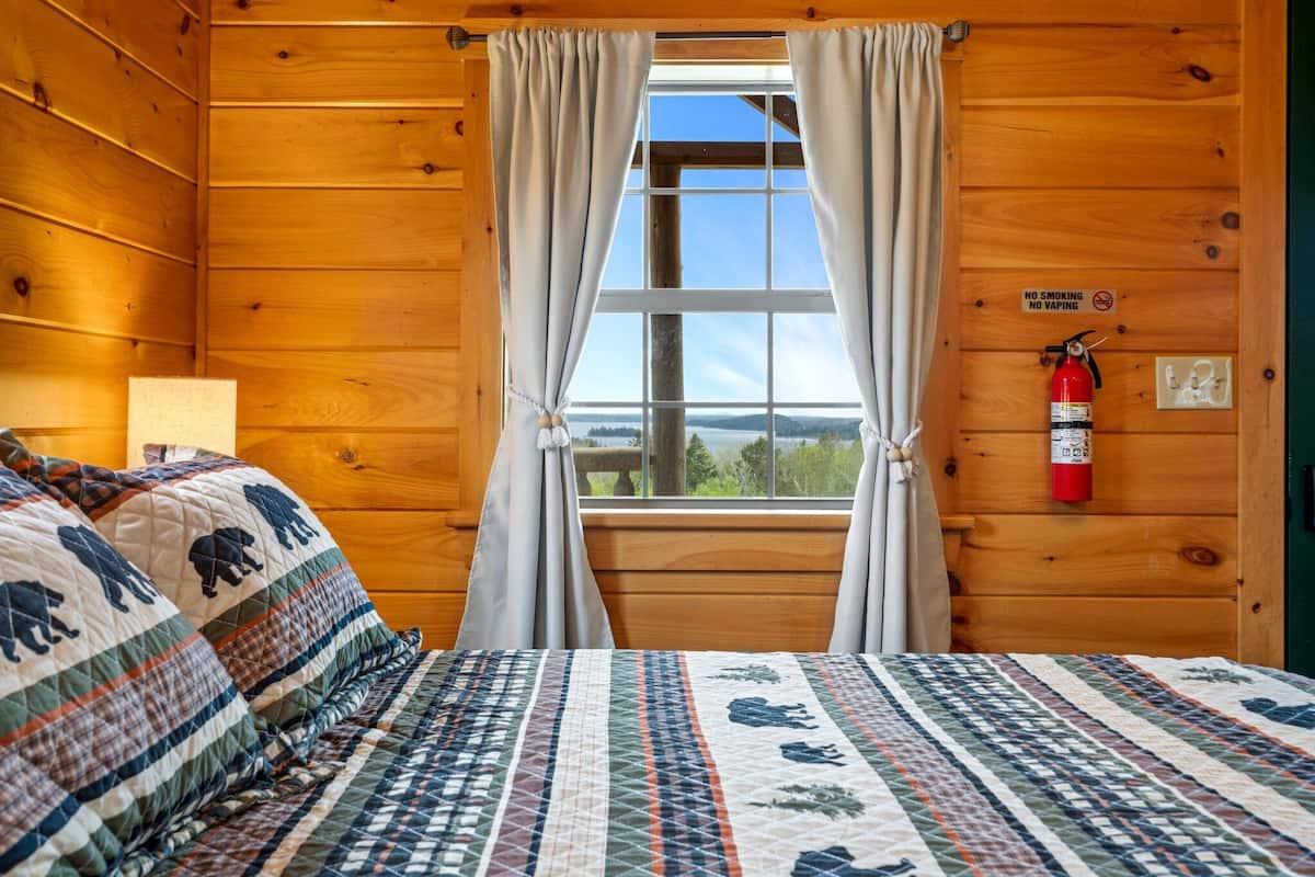 Una habitación de cabaña de madera con una cama, una ventana con cortinas, un extintor y un cartel de prohibido fumar.