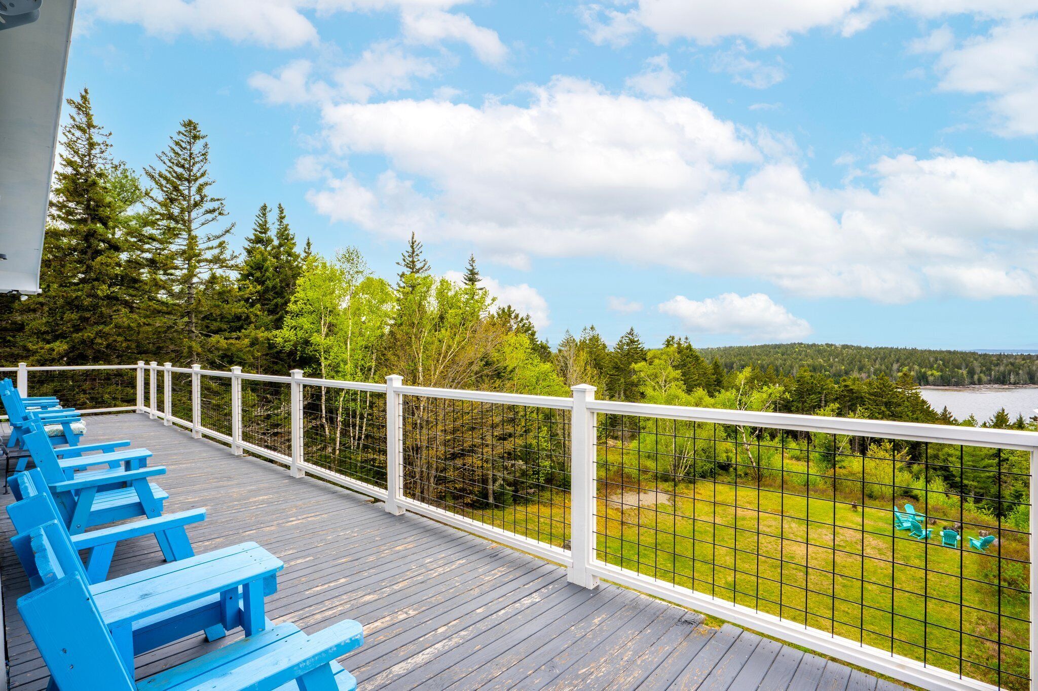 Eine Holzterrasse mit blauen Bänken und einem weißen Geländer, das auf eine Wiese mit Bäumen und einem Gewässer blickt.