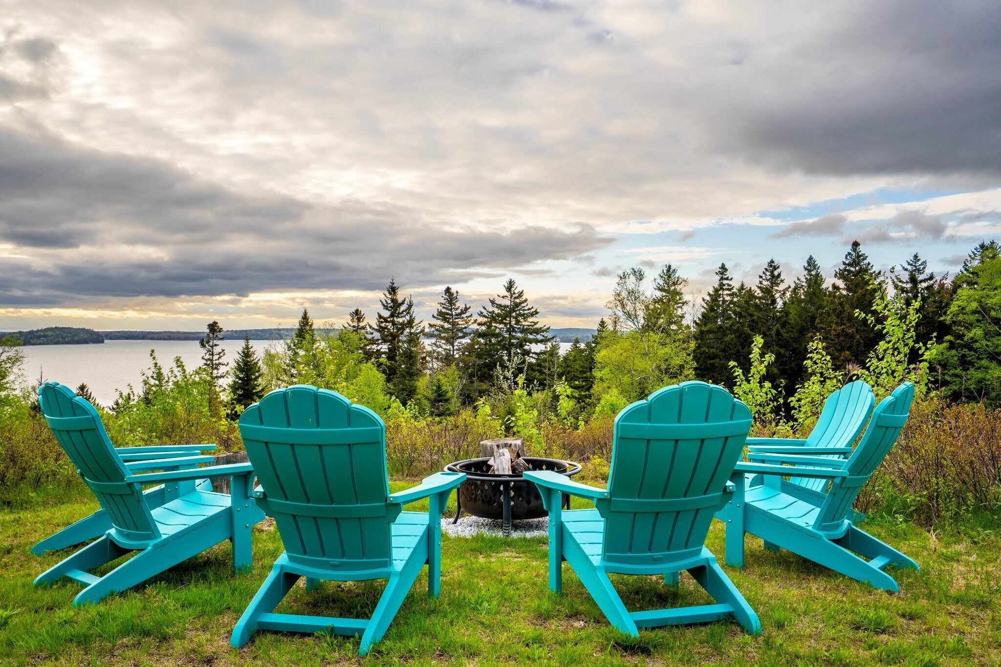 Three turquoise Adirondack chairs facing a fire pit with a lake and forest in the background.