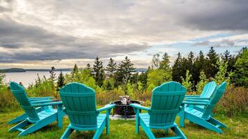 Three turquoise Adirondack chairs facing a fire pit with a lake and forest in the background.