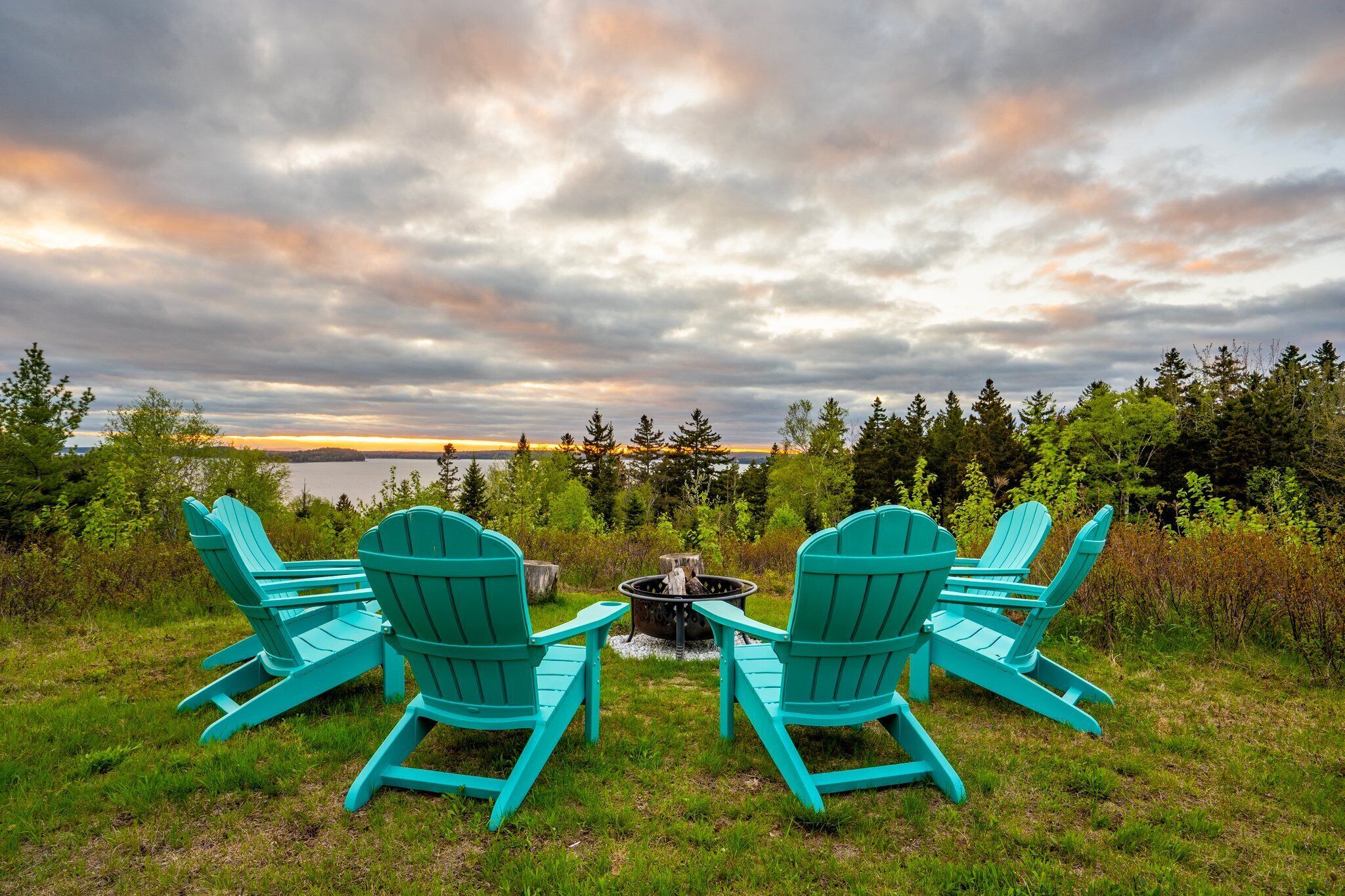 Twee turquoise Adirondack-stoelen gericht op een vuurkuil, met een meer en bos op de achtergrond.