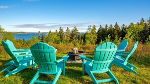 Two turquoise Adirondack chairs facing a fire pit with a view of a lake and forest.