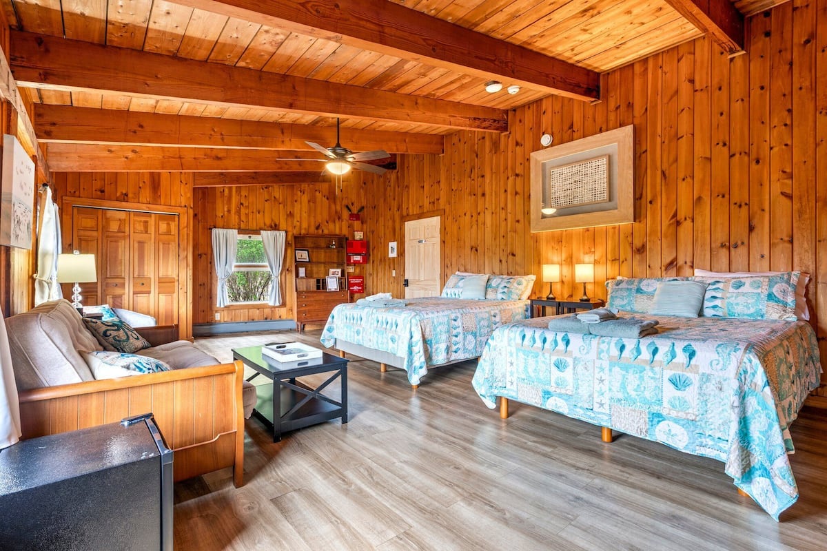 Una habitación estilo cabaña con paredes y suelos de madera, dos camas con ropa de cama azul y blanca a juego, un sofá de madera, una mesita y un ventilador de techo.