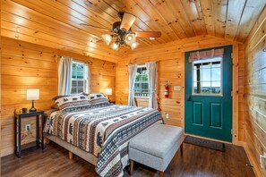 A wooden cabin bedroom with a bed, a bench, a nightstand, a ceiling fan, and a green door.