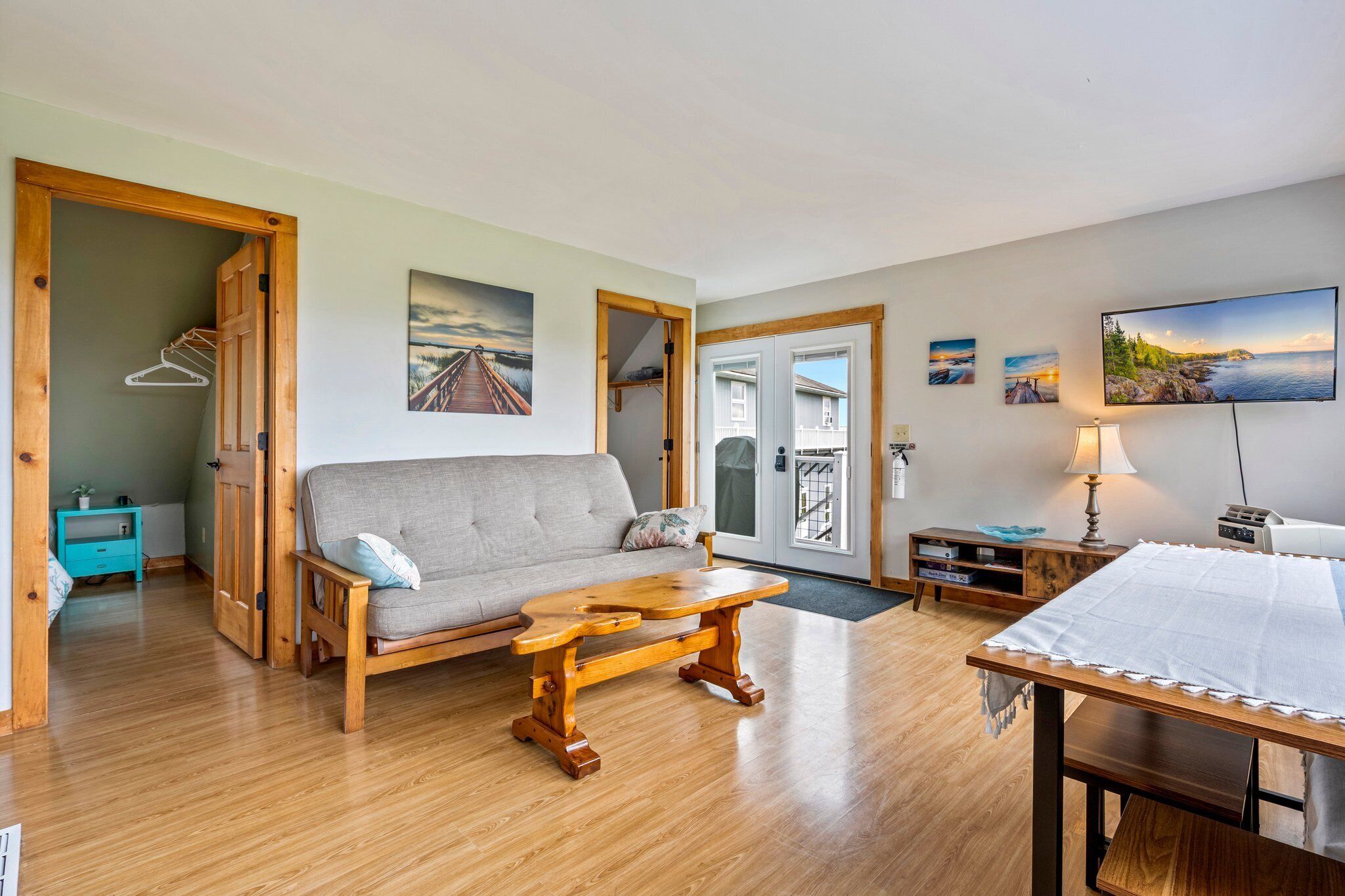 A cozy living room with a wooden bench, a sofa, and a dining table. There are doors leading to other rooms, a small table with a lamp, and a wall-mounted TV.