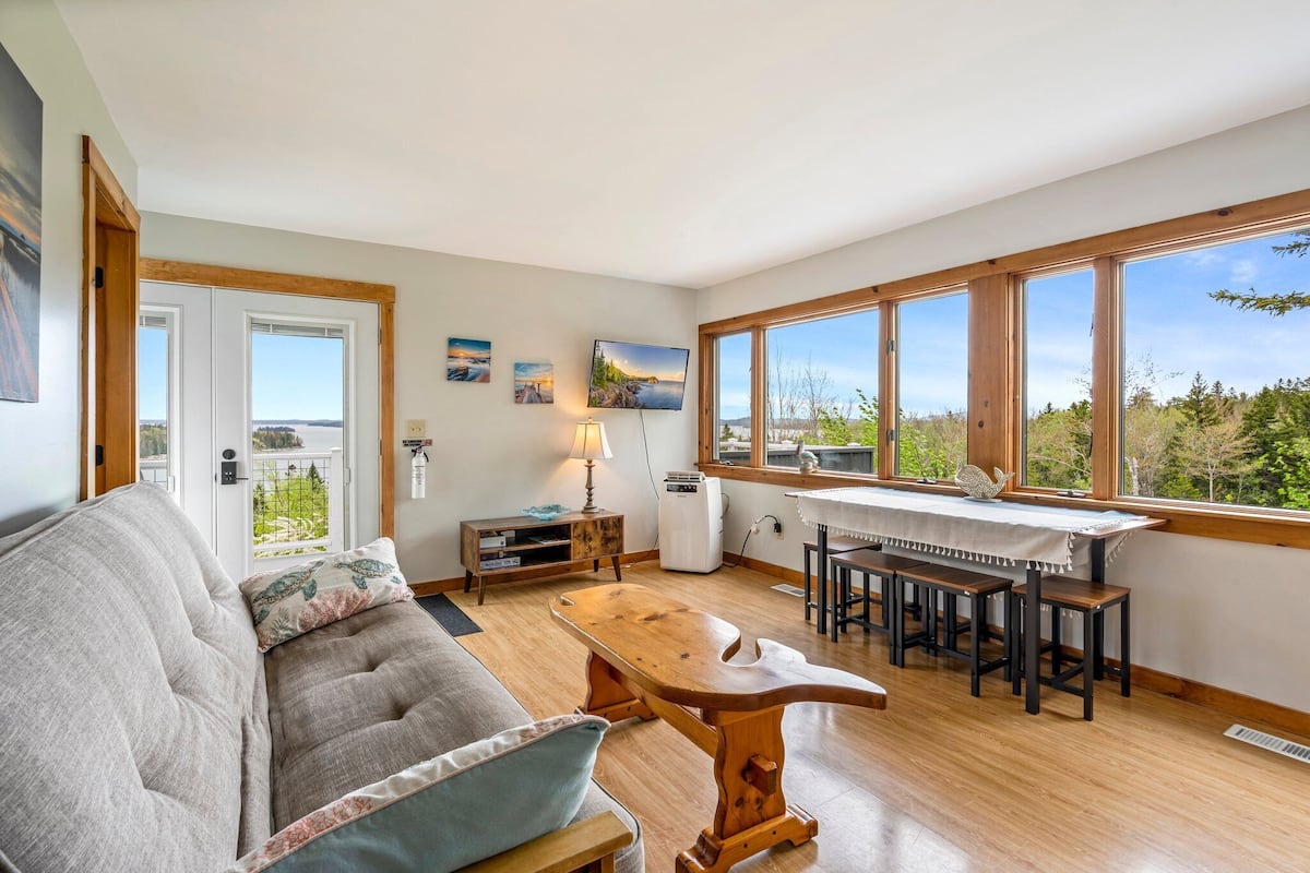 Una sala de estar acogedora con un sofá, una mesa de centro de madera y un comedor con mesa y sillas. Amplias ventanas ofrecen vistas al exterior.