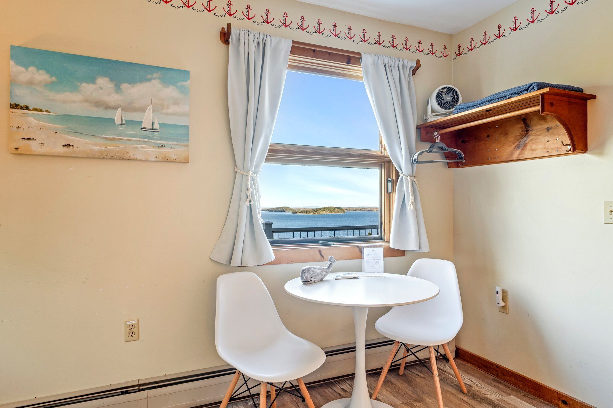 Quarto com janela, pintura de barcos à vela, mesa com duas cadeiras e prateleira de madeira com um ventilador.
