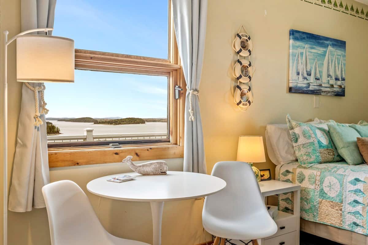 Habitación con vista a un cuerpo de agua, una mesa redonda, dos sillas, una cama con colcha y una pared con un cuadro.