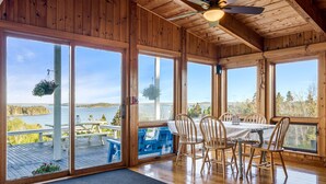 A wooden interior with large windows overlooking a lake, a dining table with chairs, and a ceiling fan.