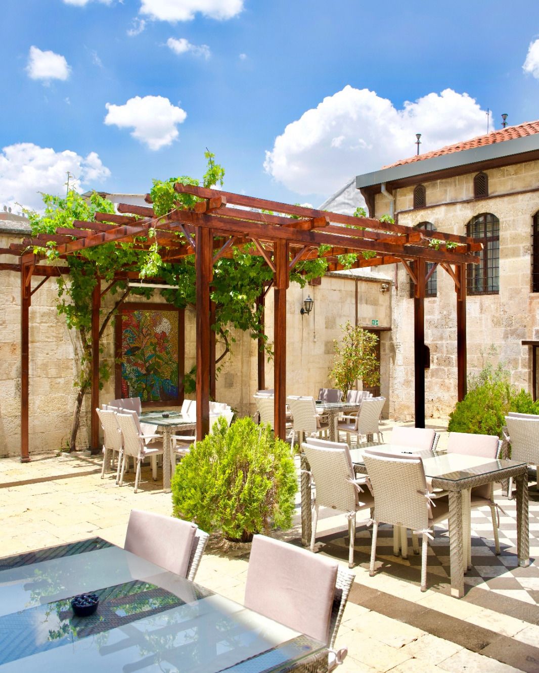Eine Terrasse im Freien mit Tischen und Stühlen, eine Pergola mit Kletterpflanzen und ein Steinhaus im Hintergrund.