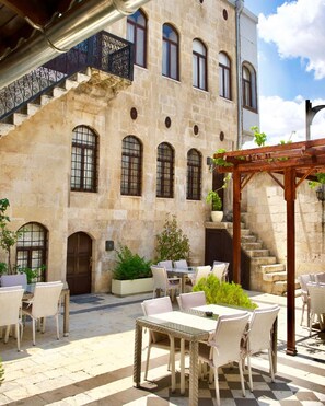 Un patio con mesas al aire libre, un edificio de piedra con ventanas de arco y una pérgola de madera.