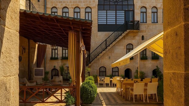 Un patio exterior con suelo de piedra, una zona de estar cubierta y un edificio grande con una ventana central arqueada y varios balcones.