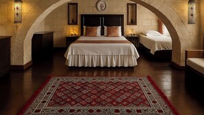 Una habitación de hotel con una cama grande, dos mesitas de noche, un tapete rojo estampado y paredes de piedra con vigas de madera.