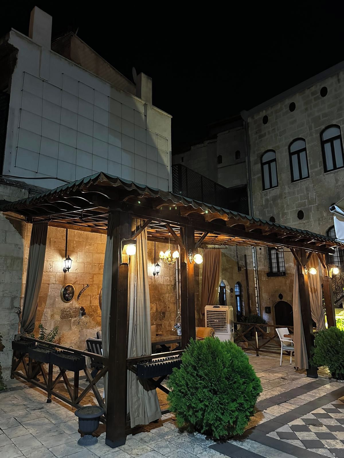 Un espace extérieur aménagé avec une pergola, des tables et des chaises, entouré de murs en pierre et d’un bâtiment doté de fenêtres en arc de cercle.