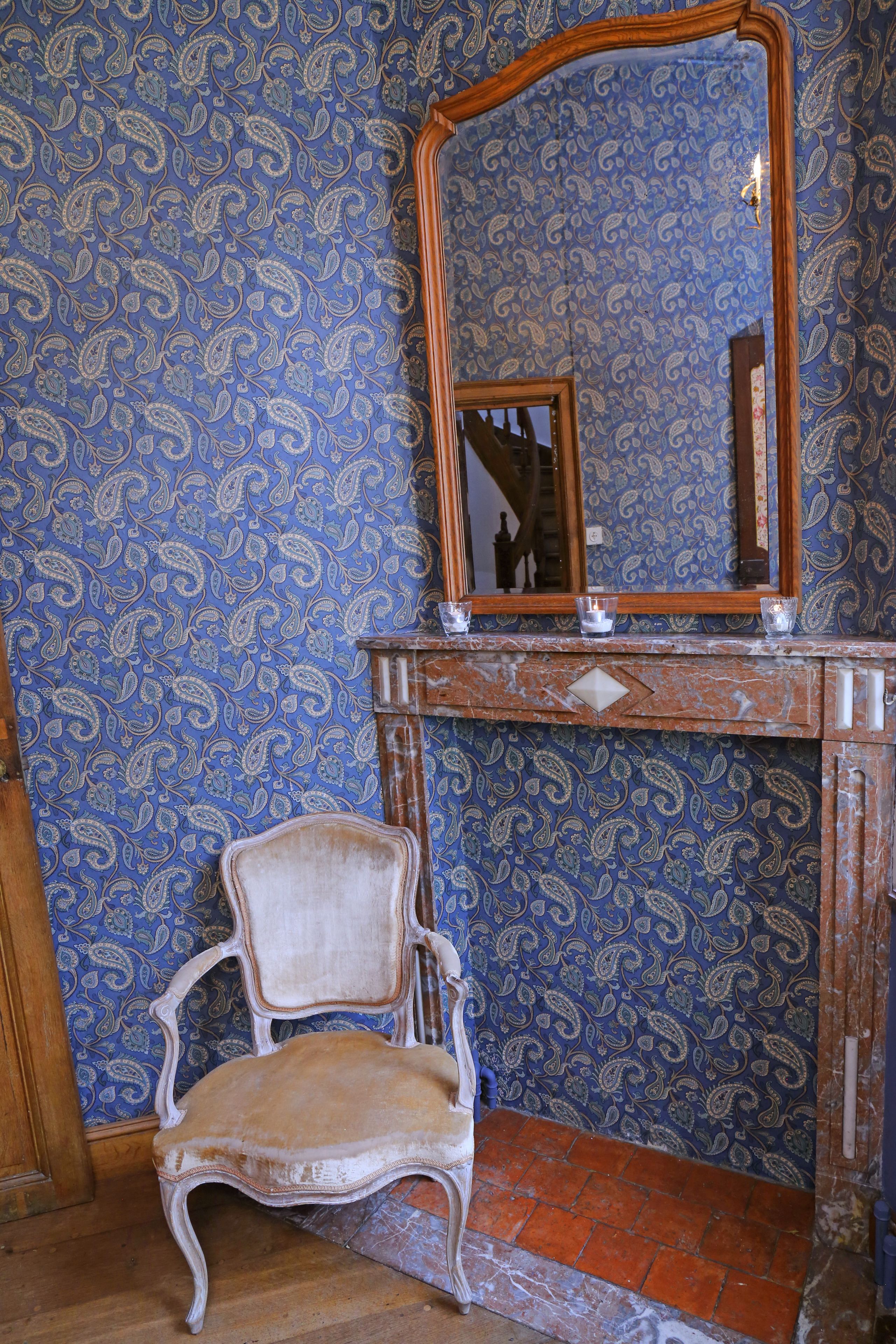 Una habitación con papel tapiz estampado, una silla de madera, un espejo y una chimenea.