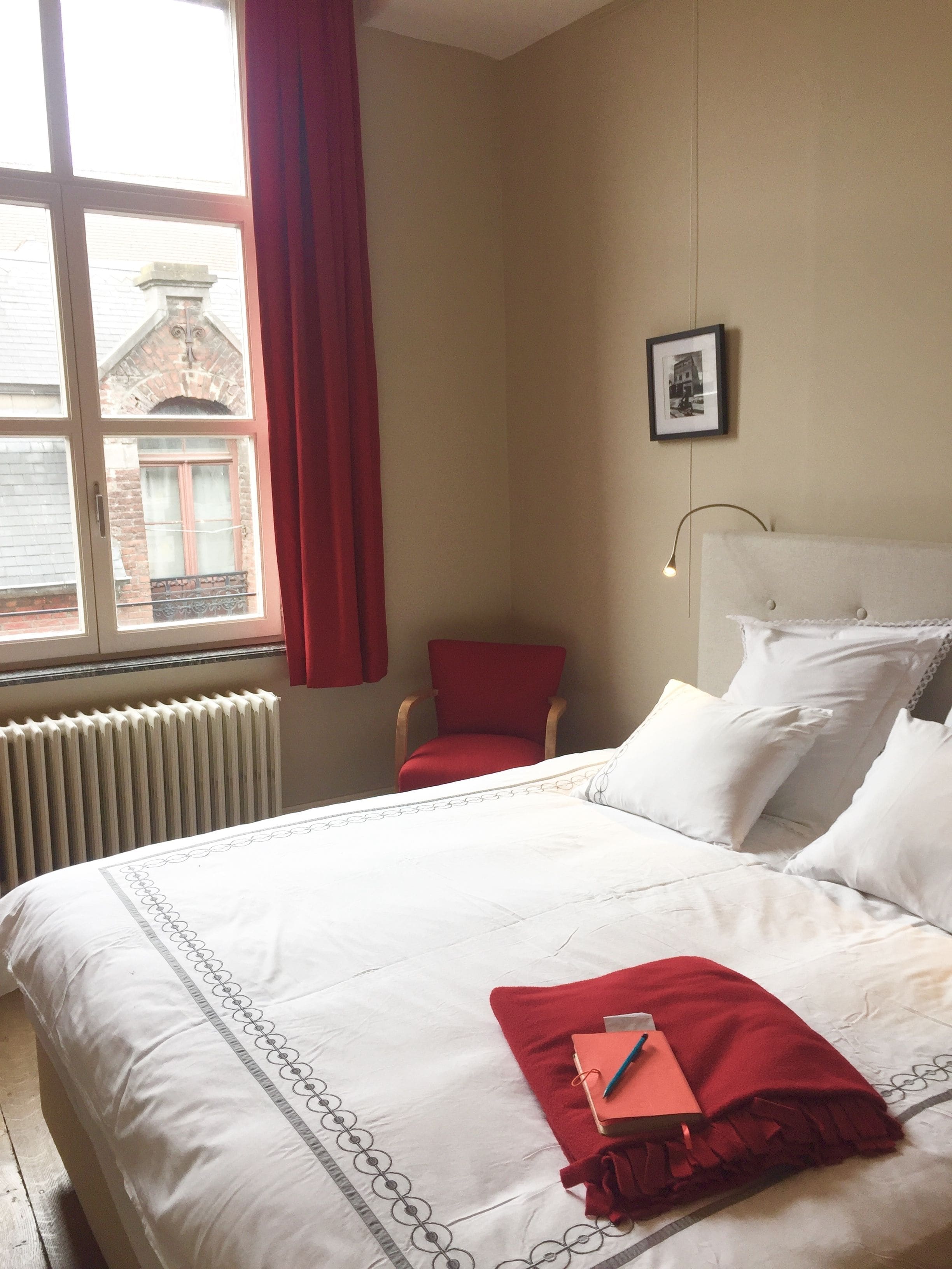 Habitación de hotel con una cama grande, una silla roja, un radiador y una ventana con vista a un edificio.
