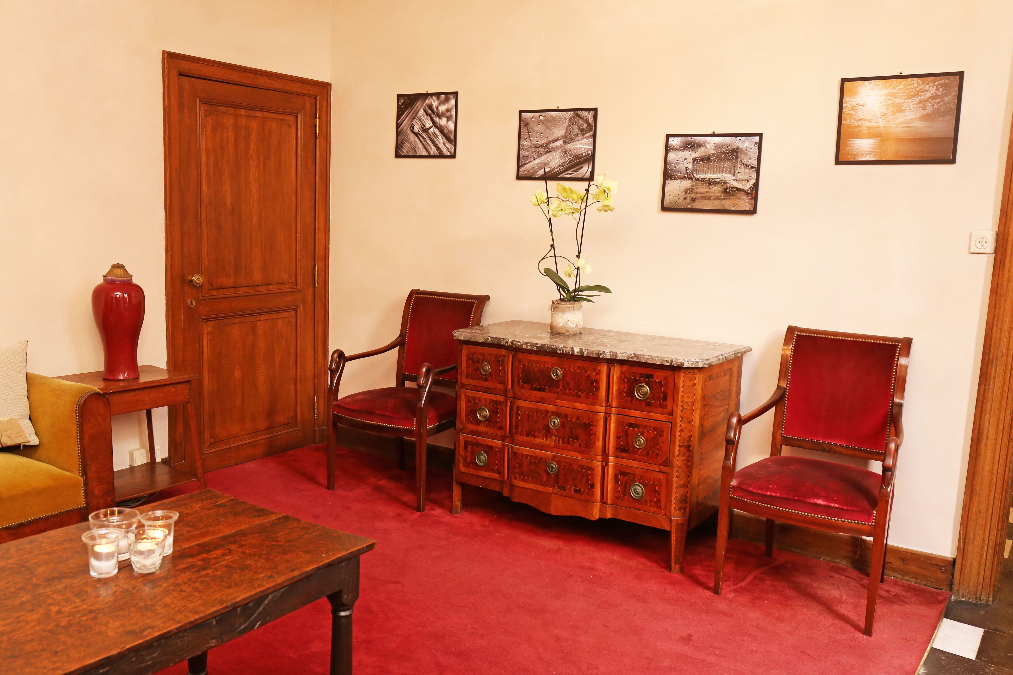A room with a wooden door, a red vase, a wooden cabinet with drawers, two red upholstered chairs, a small table with candles, and three framed pictures on the wall.
