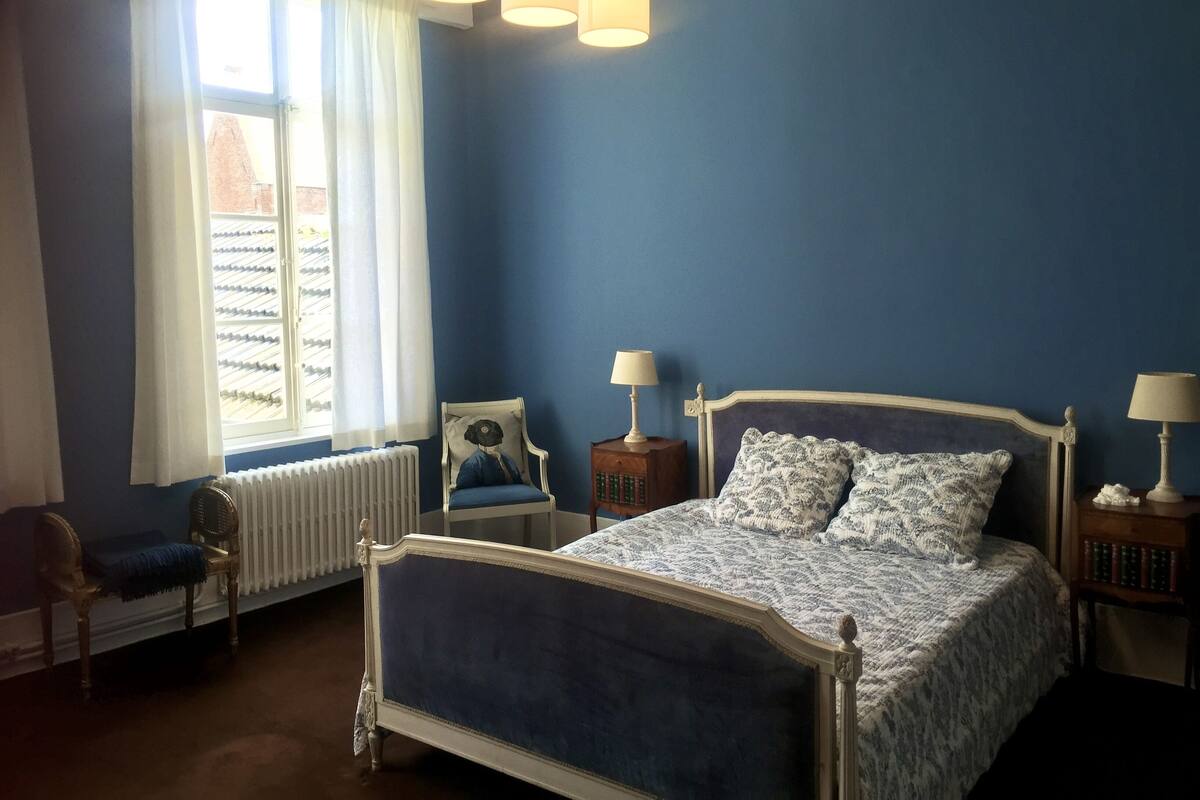Ein Schlafzimmer mit einem großen Bett, einem Fenster mit Vorhängen, einem Heizkörper, einem Sessel und zwei Nachttischen mit Lampen.