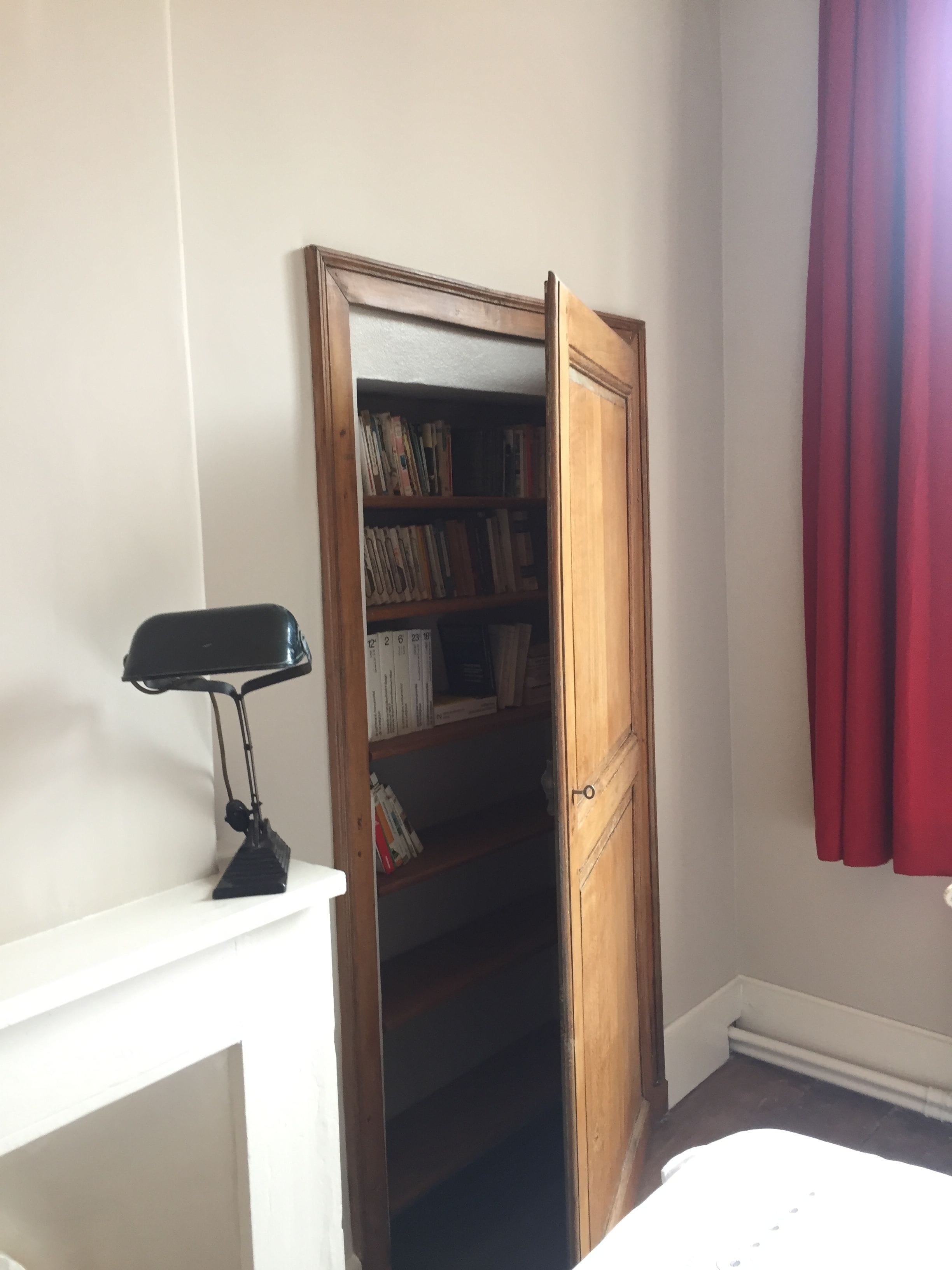 A room with a wooden door leading to a bookshelf, a desk with a lamp, and a red curtain.