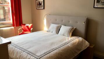 Een net opgemaakt bed met een witte deken en decoratieve randen, een rode stoel, een nachtkastje en een ingelijste foto aan de muur.