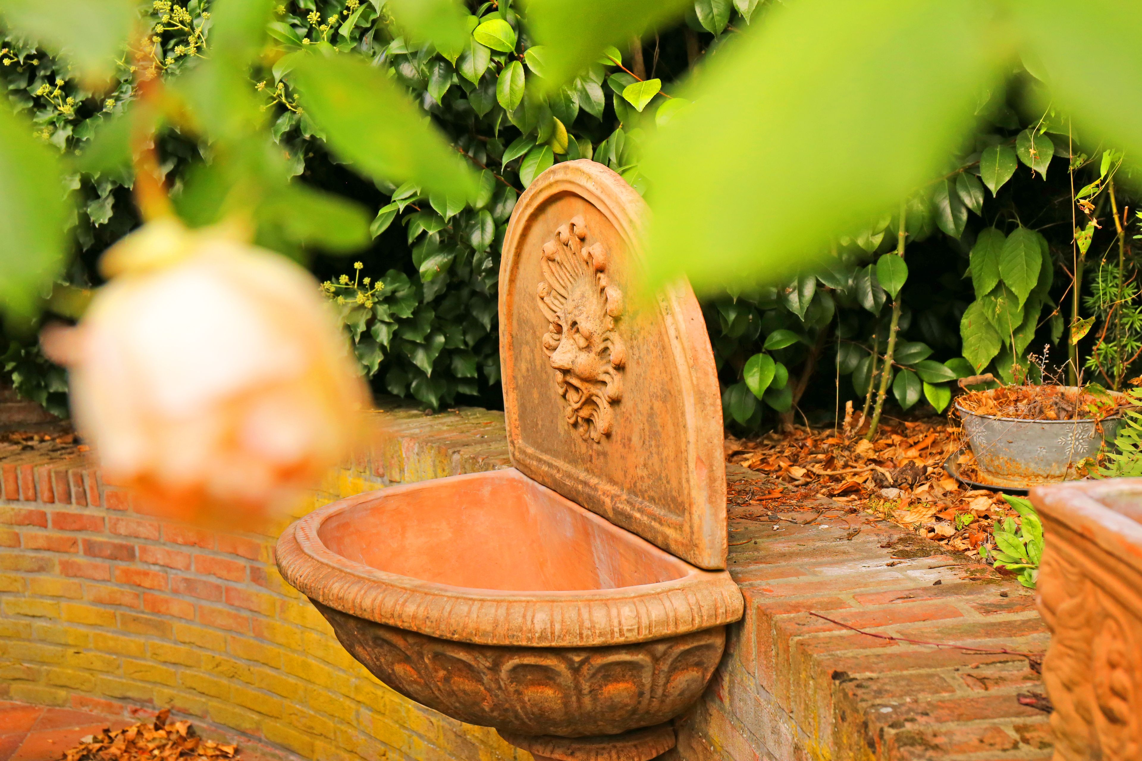 A terracotta fountain with a decorative lid, surrounded by lush greenery and a brick wall.