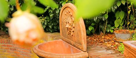 A terracotta fountain with a decorative lid, surrounded by lush greenery and a brick wall.