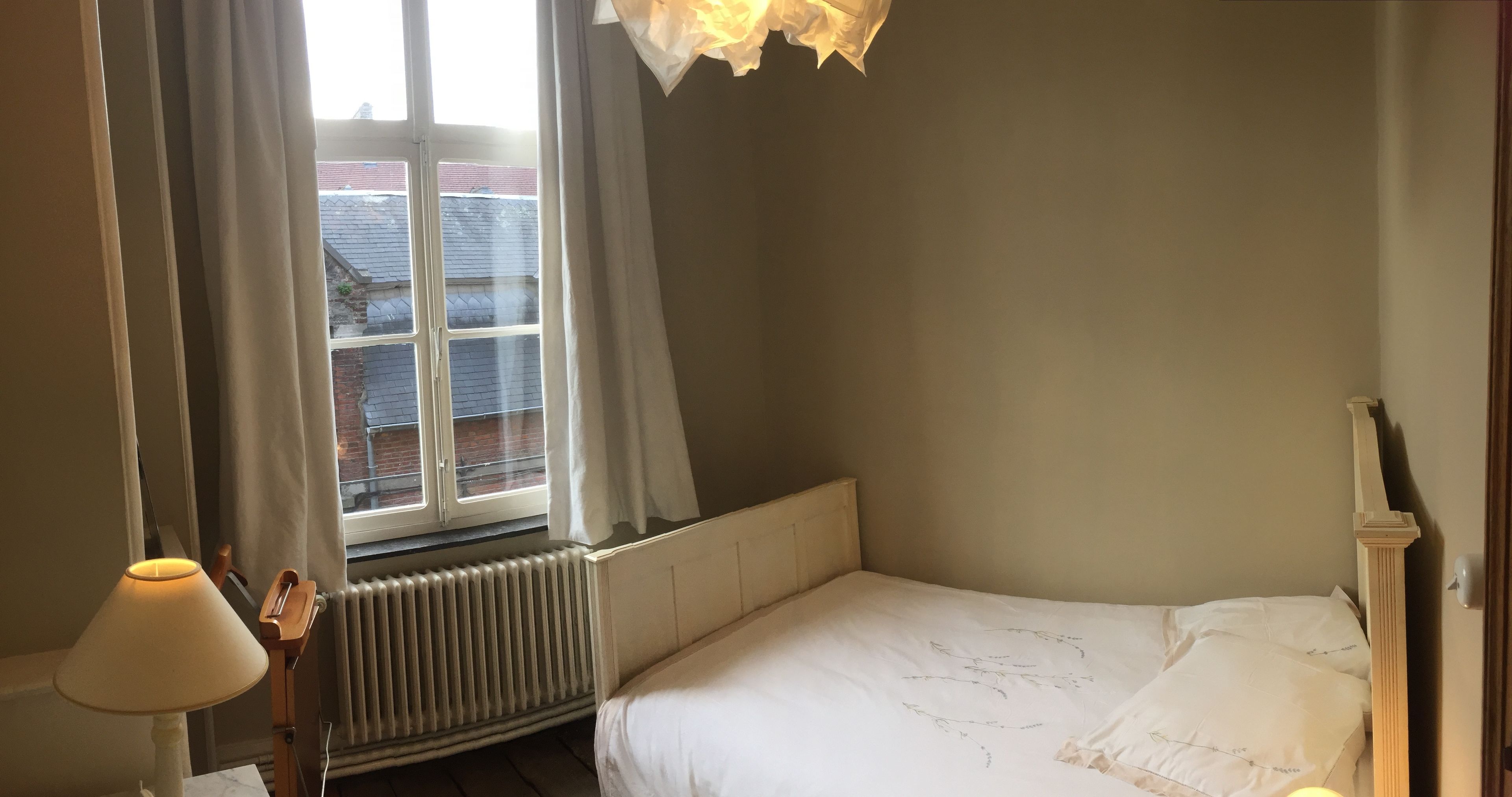 A bedroom with a single bed, a radiator, a window with curtains, and a lamp.